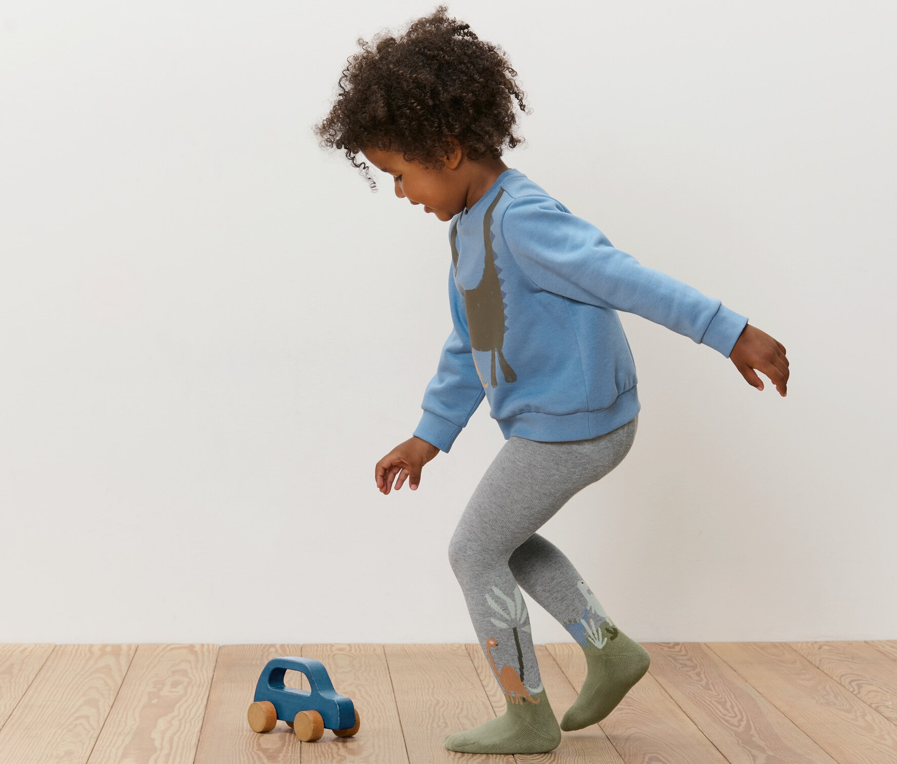 Dziecko bawi się niebieskim drewnianym autkiem, ma na sobie niebieską bluzę i szare rajstopy. Rajstopy dziecięce, 3 pary, niebieskie.