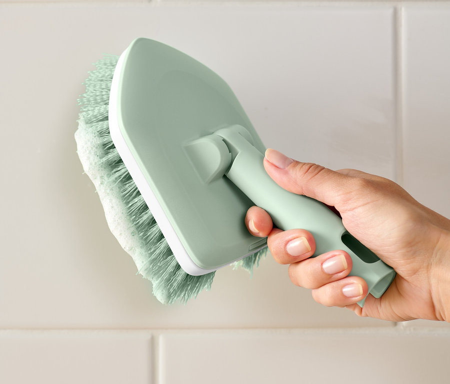 Ręka trzyma szczotkę do szorowania płytek i wanien i czyści białe płytki.