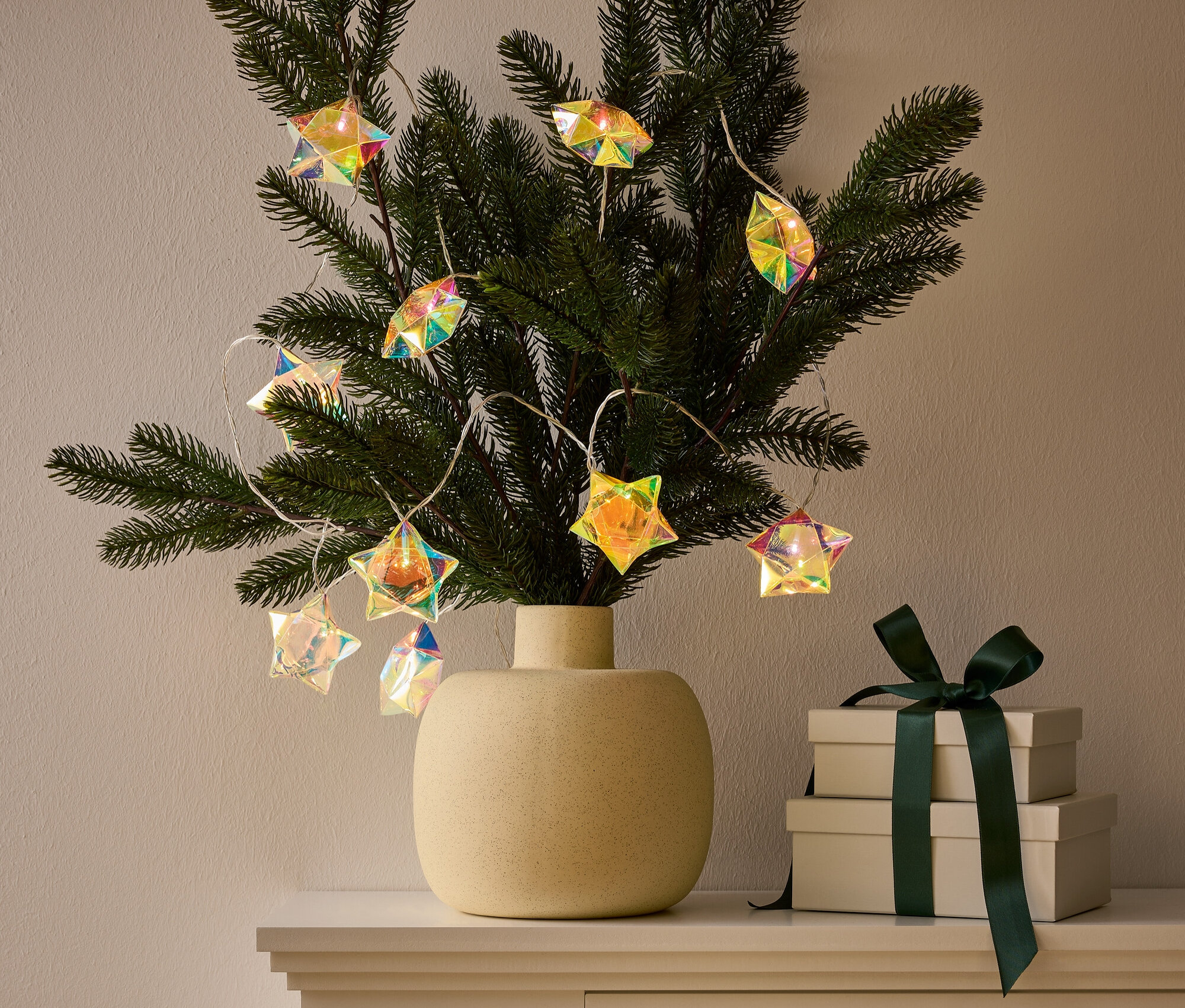 Wazon z gałązką ozdobioną girlandą świetlną w kształcie gwiazdy stoi na szafce obok dwóch pudełek prezentowych z ciemnozieloną wstążką.