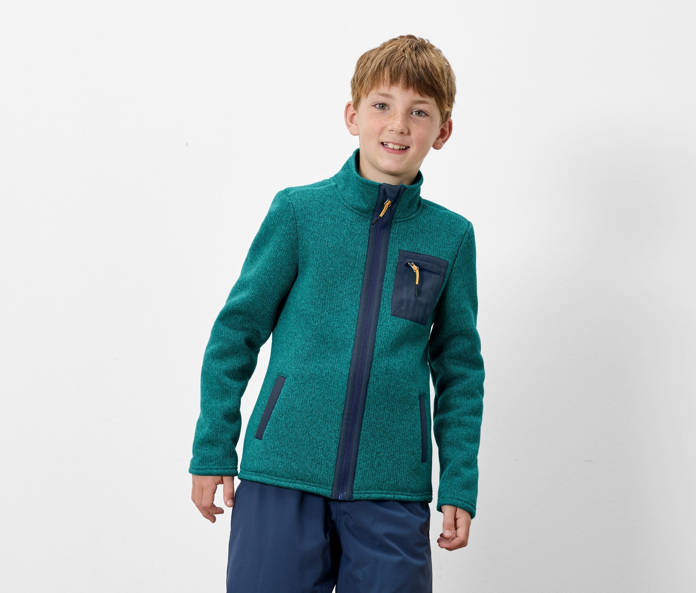 Chłopiec ma na sobie zieloną bluzę polarową na zamek z granatowymi detalami i niebieskie spodnie.