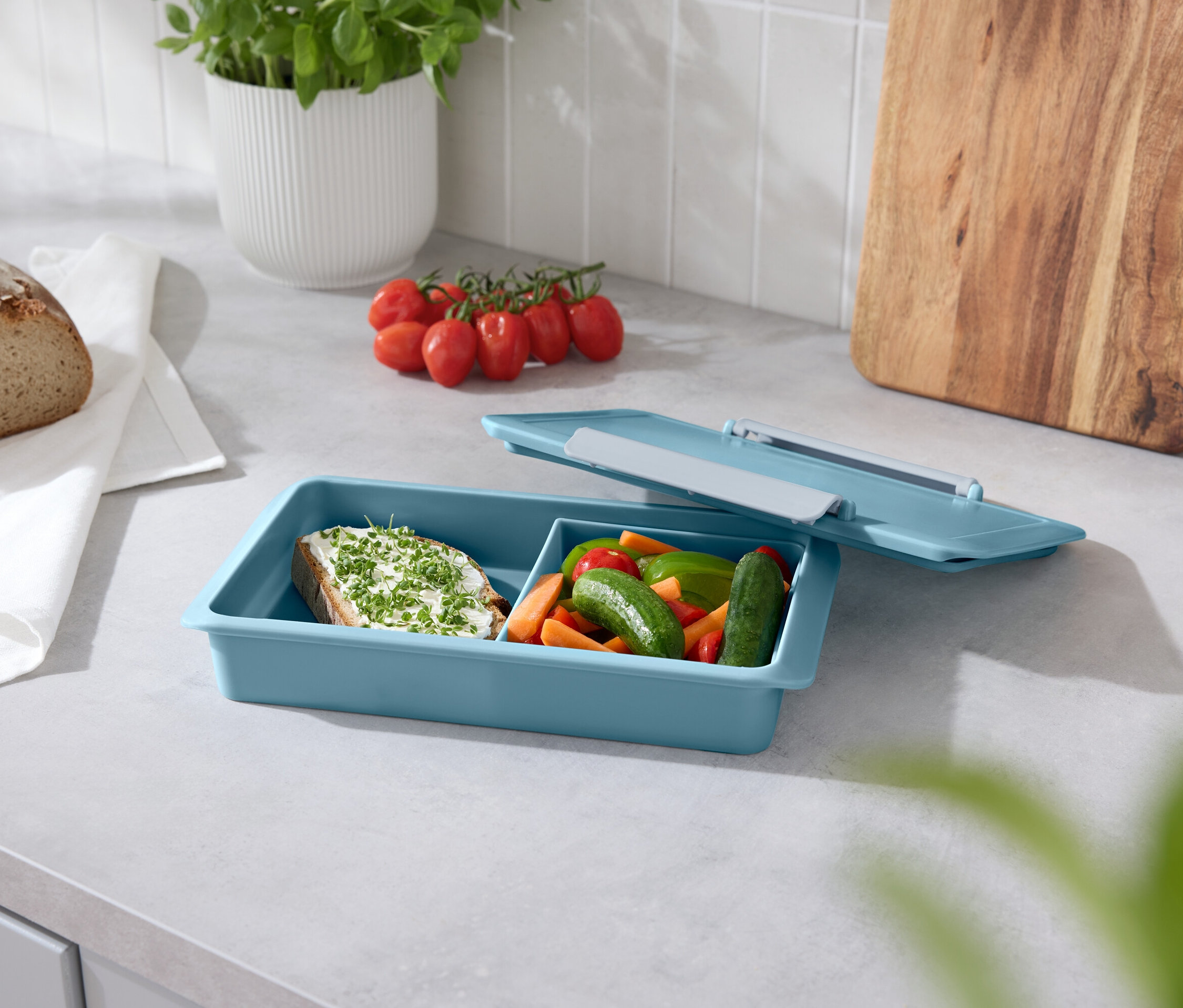 Chleb, pomidory, bazylia i niebieskie pudełko na przekąski z otwartą pokrywką na blacie.