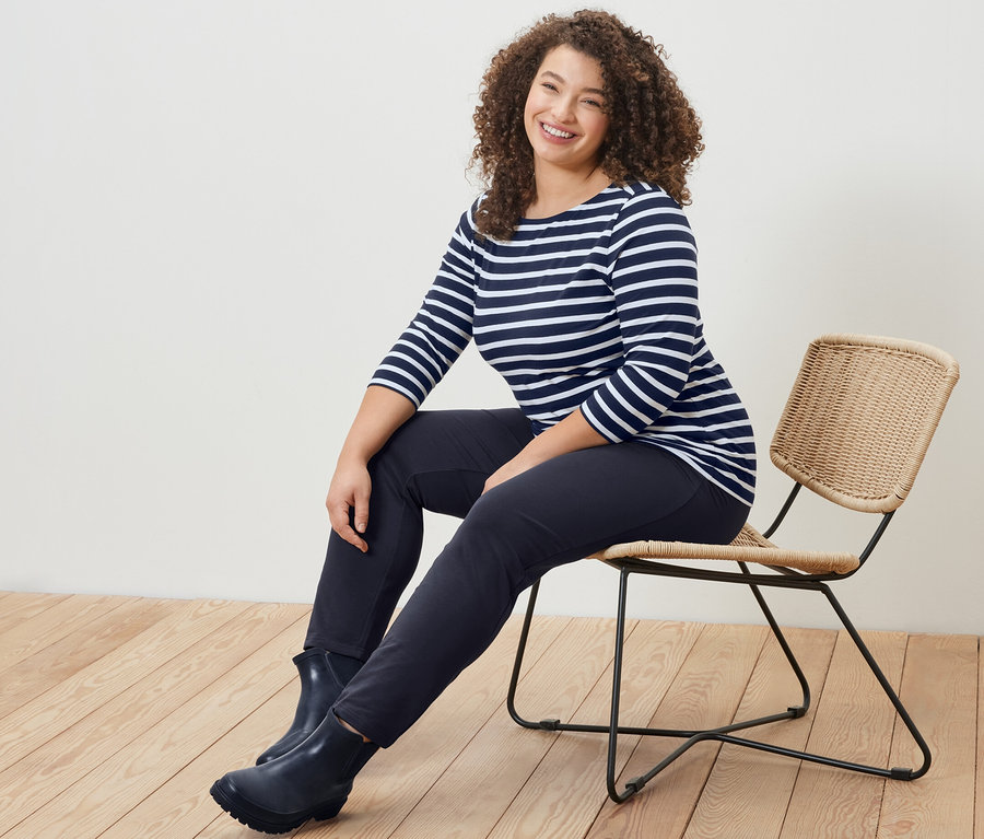 Kobieta siedzi na krześle w bluzce w paski z rękawami o długości 3/4 i granatowych spodniach dresowych.