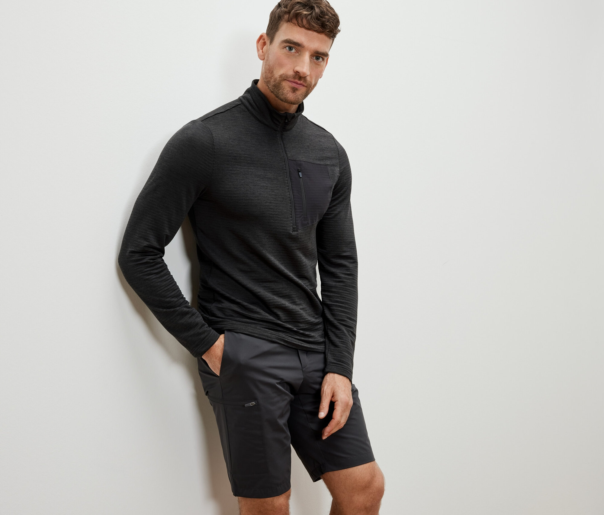 Mężczyzna oparty o ścianę ma na sobie antracytową koszulkę funkcyjną z długim rękawem i szorty.