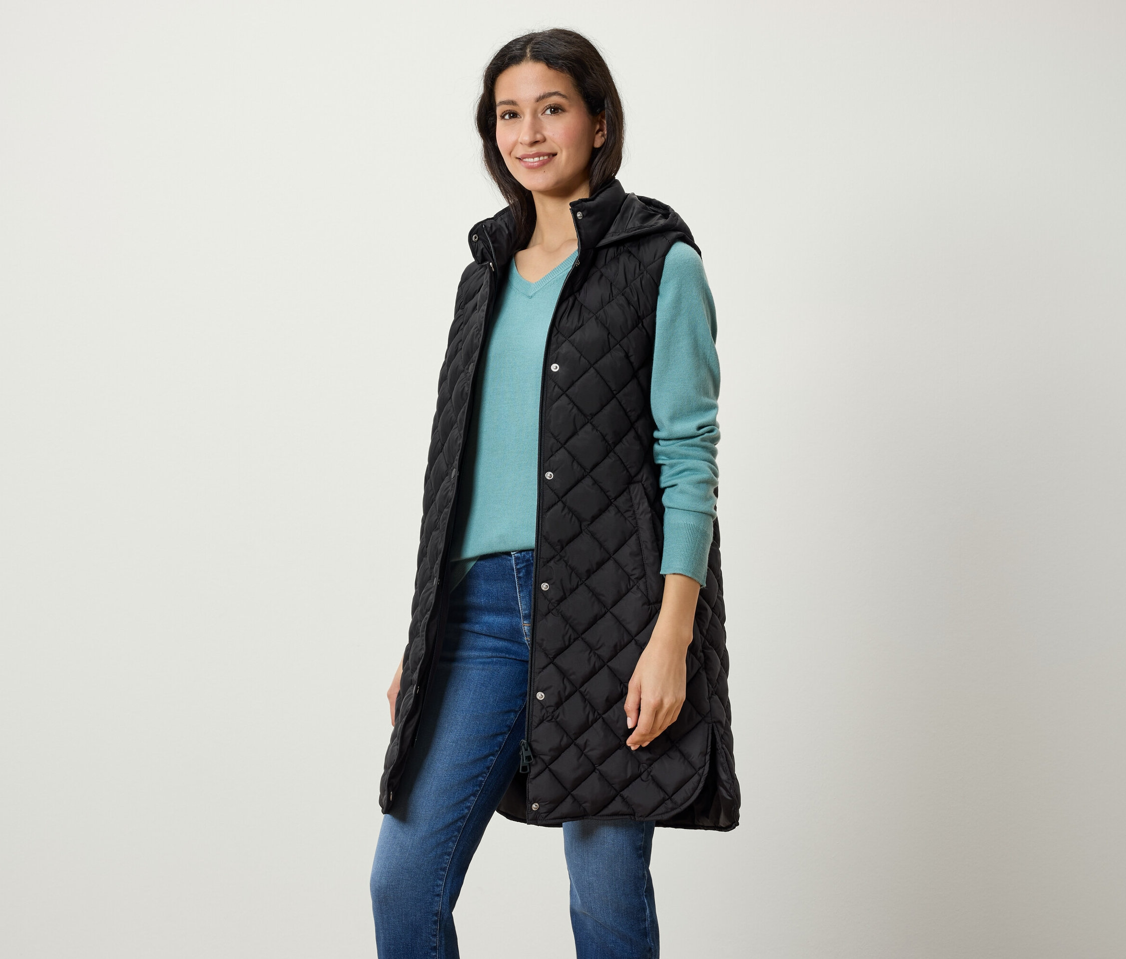 Kobieta ma na sobie długą, czarną, pikowaną kamizelkę z kapturem, niebieski sweter i dżinsy.