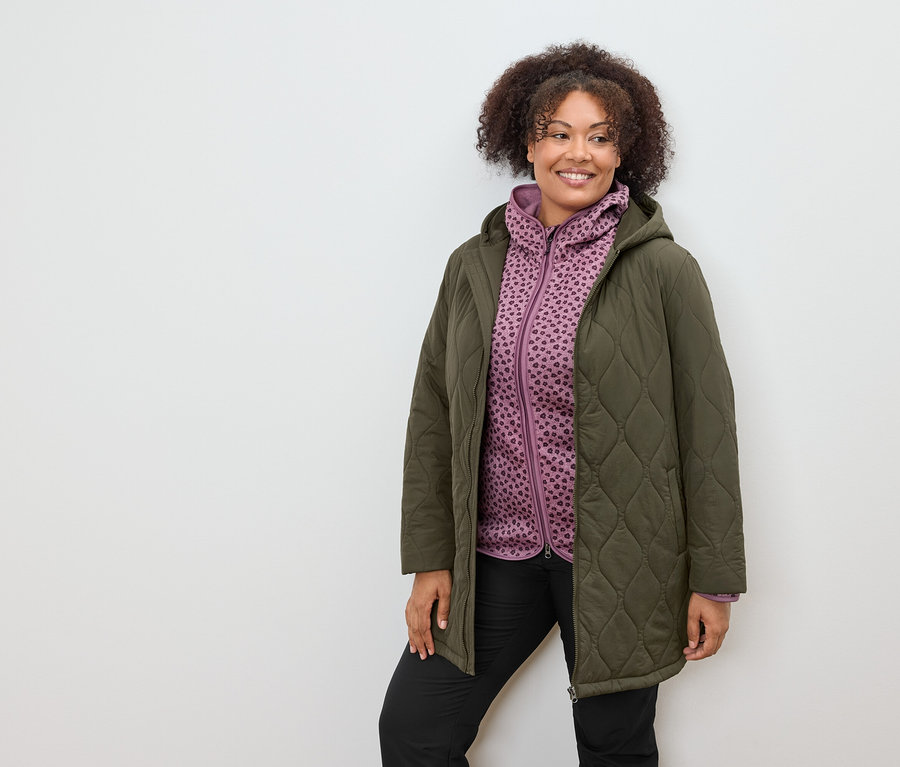 Kobieta w oliwkowym płaszczu i fioletowej bluzie z kapturem w wzory.