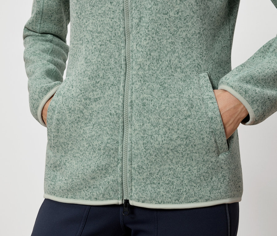 Osoba trzyma ręce w kieszeniach zielonej kurtki polarowej.