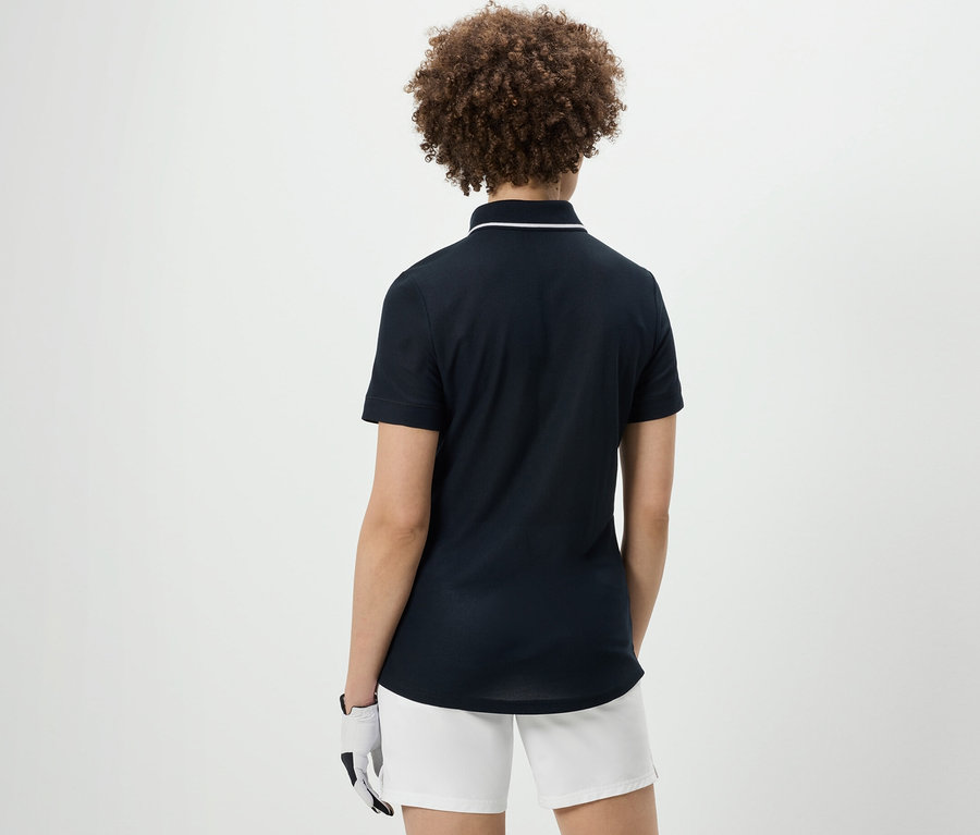 Widok z tyłu kobiety z kręconymi włosami w granatowej funkcyjnej koszulce polo i białych szortach.