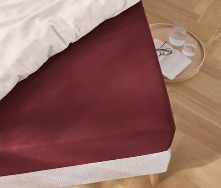 Zbliżenie czerwonego prześcieradła na łóżku, obok którego stoi okrągły stolik z okularami, książką i szklanką wody.