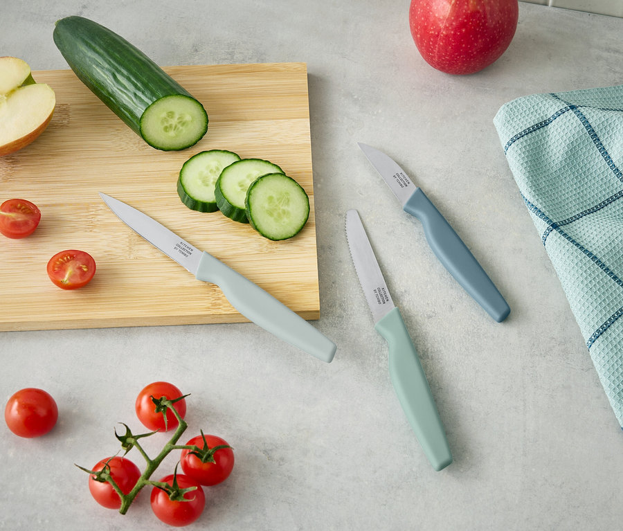 Na drewnianej desce leży połowa jabłka, ogórek i pokrojone plasterki ogórka. Obok leżą trzy noże i pomidorki cherry.