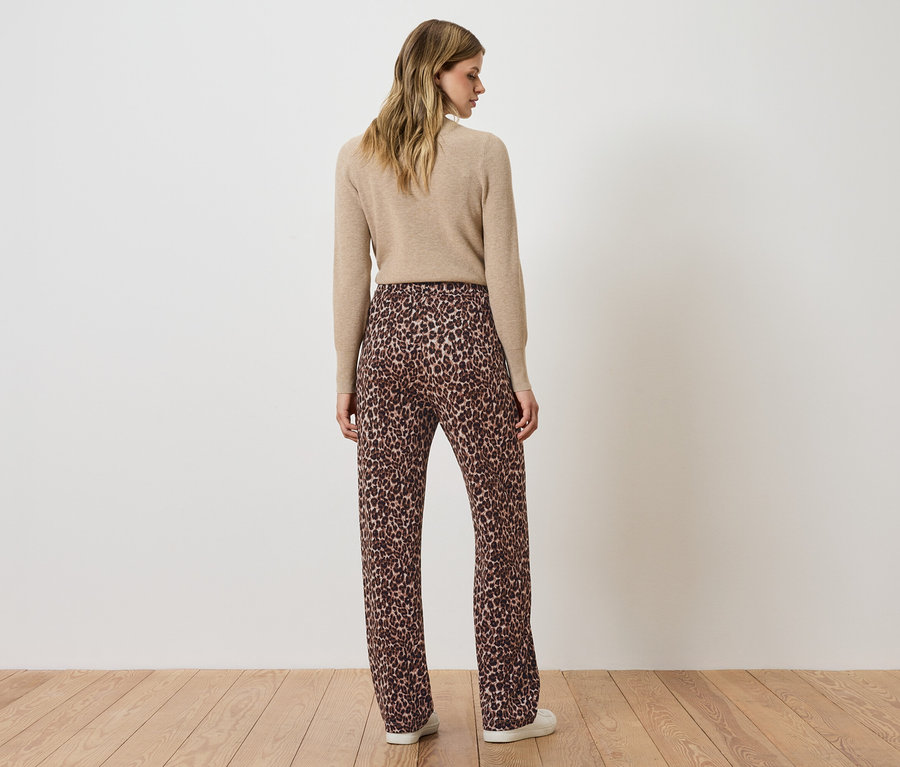 Kobieta stoi tyłem do aparatu, ubrana w beżowy sweter i brązowe spodnie we wzór zwierzęcy.