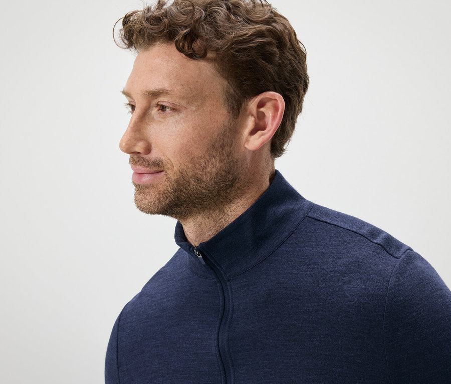 Portret mężczyzny z brązowymi, falowanymi włosami i brodą, ubranego w niebieski sweter zapinany na zamek.