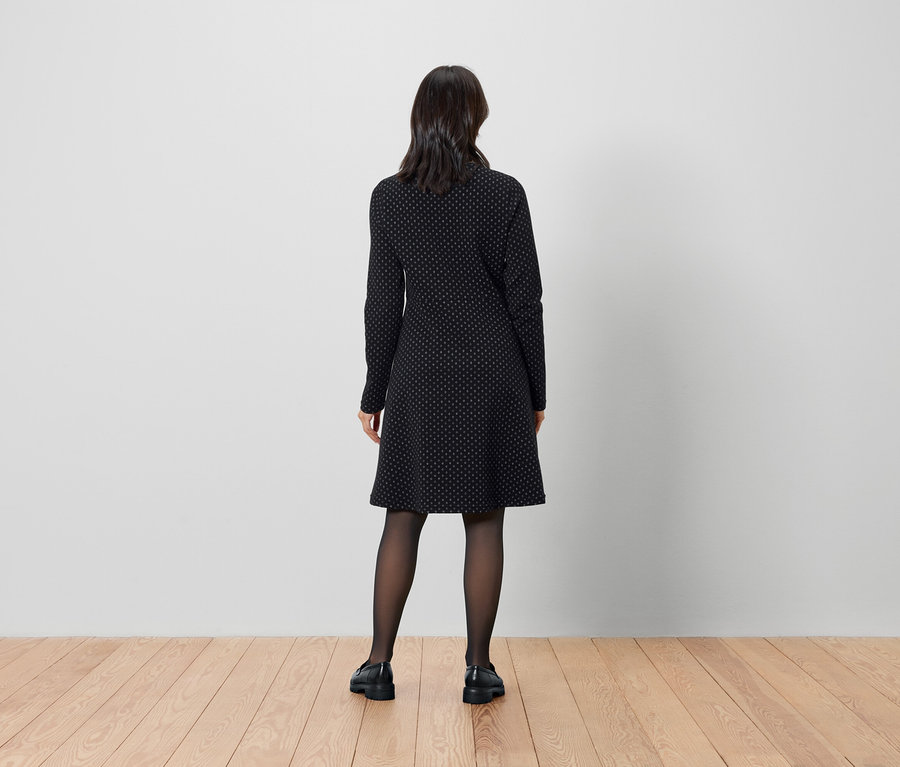 Kobieta stoi tyłem do aparatu, ubrana w czarną sukienkę i rajstopy.