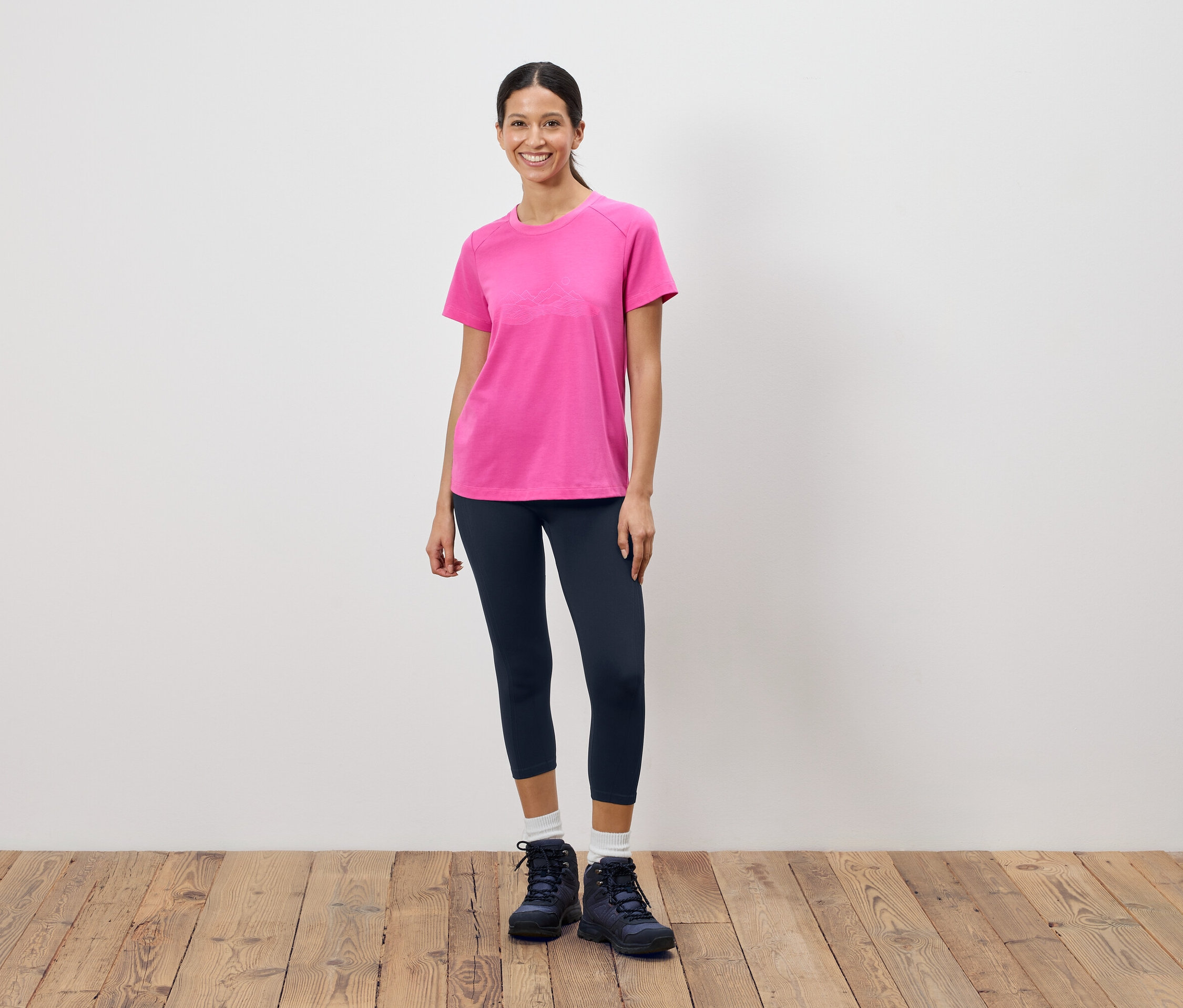 Kobieta stoi na tle białej ściany, ubrana w różowy T-shirt, ciemne legginsy i buty trekkingowe.