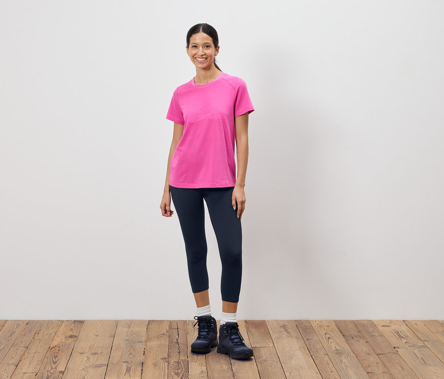 Kobieta stoi na tle białej ściany, ubrana w różowy T-shirt, ciemne legginsy i buty trekkingowe.