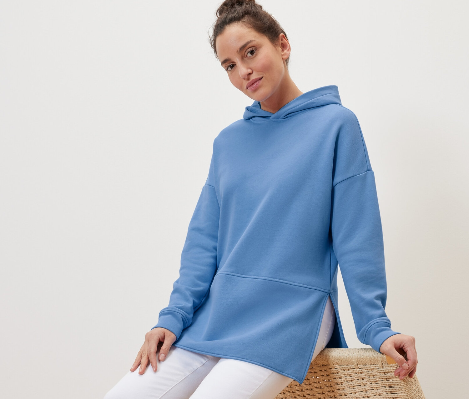 Kobieta siedzi na wiklinowym krześle, ubrana w jasnoniebieską, długą bluzę z kapturem.