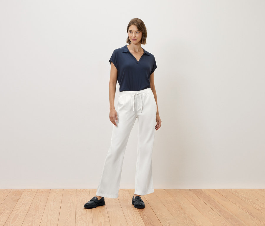 Kobieta stoi na drewnianej podłodze, ubrana w granatowy t-shirt i białe spodnie.