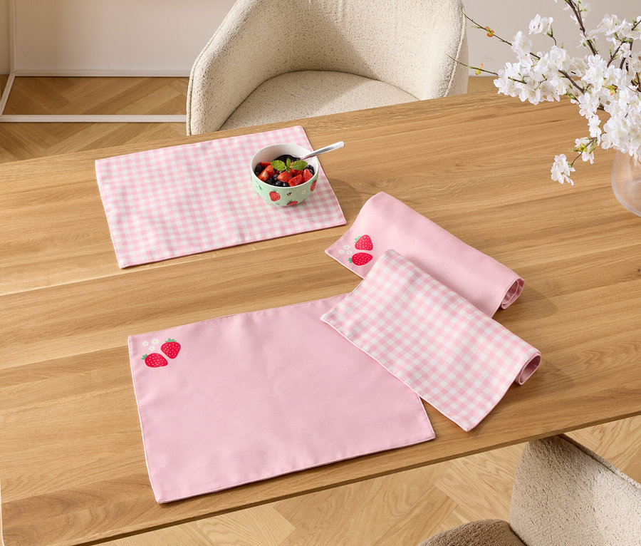 Na drewnianym stole leżą różowe podkładki, jedna w truskawki, miseczka z jagodami i wazon z kwitnącymi gałązkami.