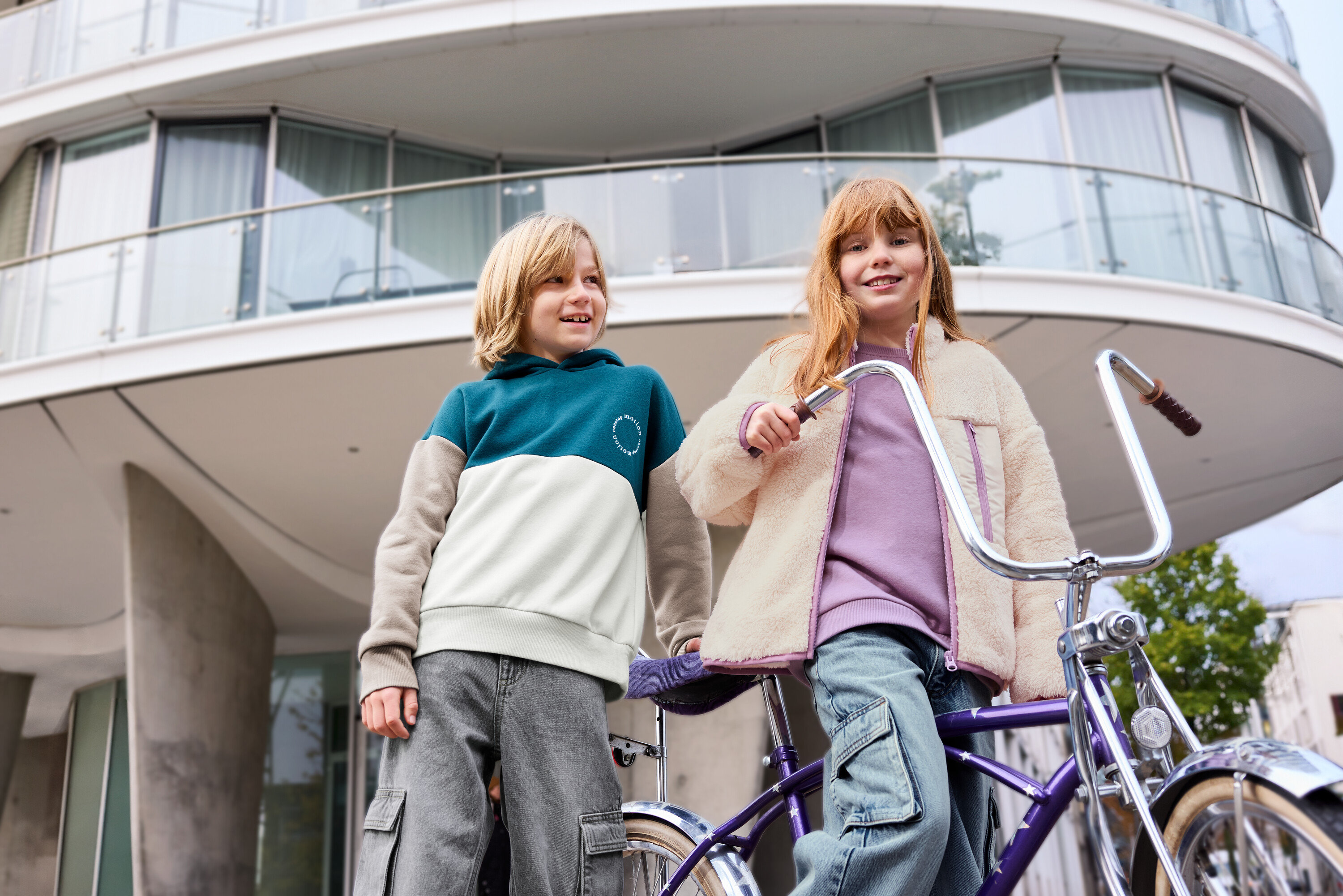 Dwoje dzieci stoi przed budynkiem, jedno dziecko ma na sobie lawendową bluzę dziecięcą z drapanym wnętrzem.