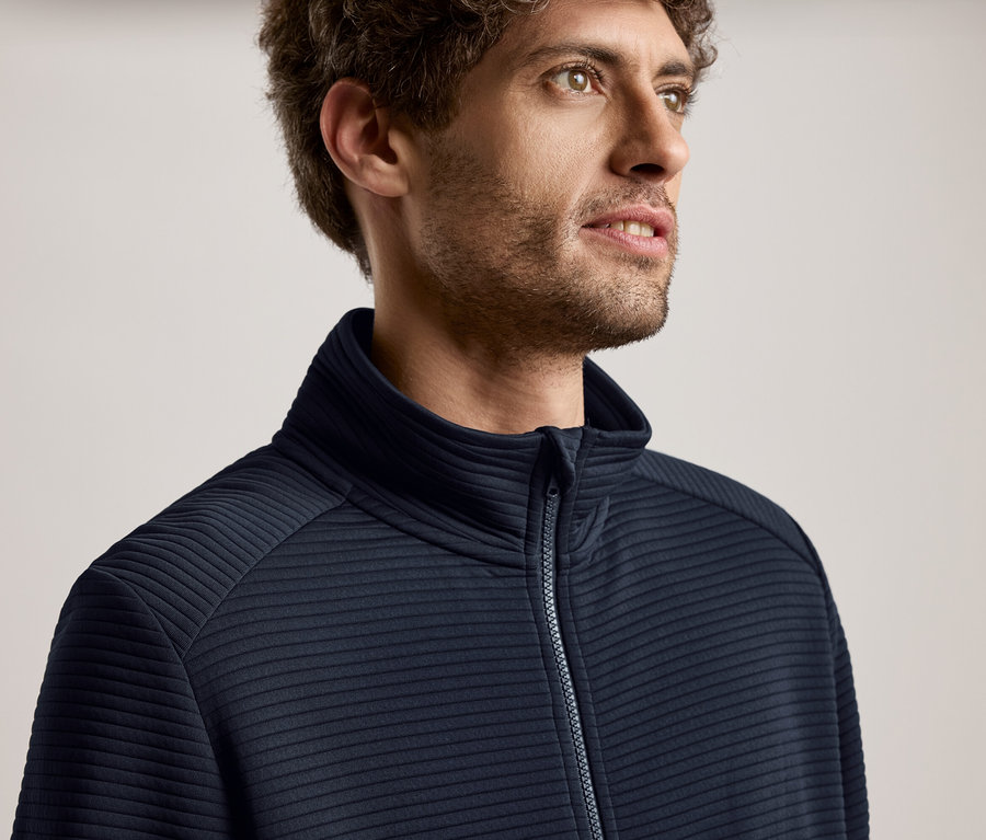 Portret mężczyzny o kręconych włosach w granatowej kurtce zapinanej na zamek, patrzącego w prawo.