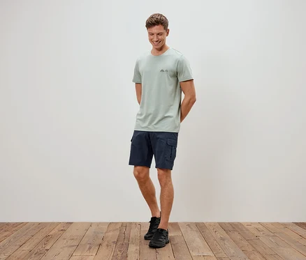 Mężczyzna stoi na tle ściany, ubrany w jasnozielony t-shirt, niebieskie szorty i czarne buty.