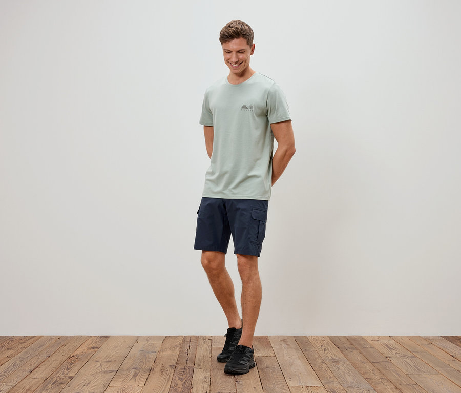 Mężczyzna stoi na tle ściany, ubrany w jasnozielony t-shirt, niebieskie szorty i czarne buty.