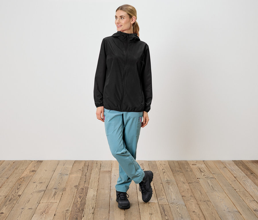 Kobieta w czarnej kurtce przeciwdeszczowej z kapturem, jasnoniebieskich spodniach i czarnych butach stoi na tle białej ściany.