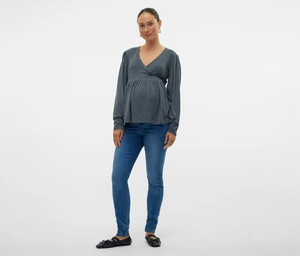 Kobieta w szarej bluzce kopertowej i dżinsach skinny MAMALICIOUS „MLMILA” pozuje na białym tle.
