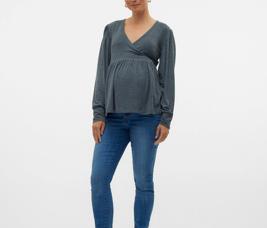 Kobieta w ciąży ma na sobie szarą bluzkę z długim rękawem, niebieskie dżinsy i czarne baleriny.