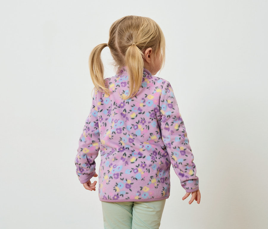 Widok z tyłu małej dziewczynki z blond warkoczami, ubranej w liliową bluzę polarową w kwiaty i zielone spodnie.