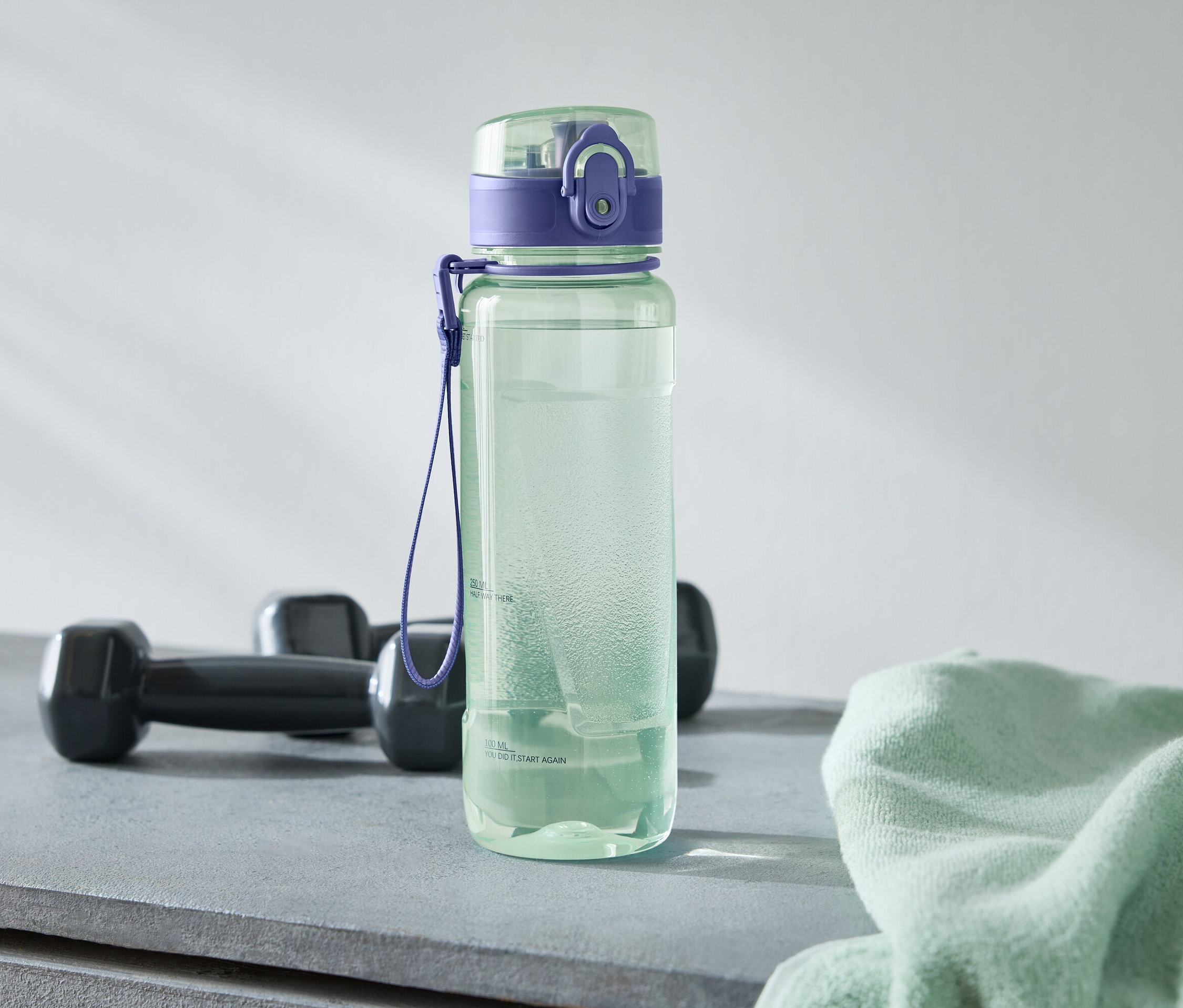 Zielona butelka na wodę z fioletową nakrętką stoi obok dwóch czarnych hantli i jasnozielonego ręcznika na szarym tle.