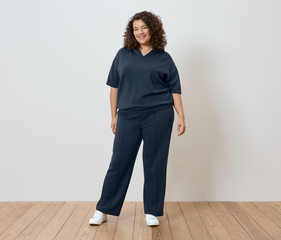 Kobieta z kręconymi włosami w granatowym T-shircie i spodniach na tle białej ściany.