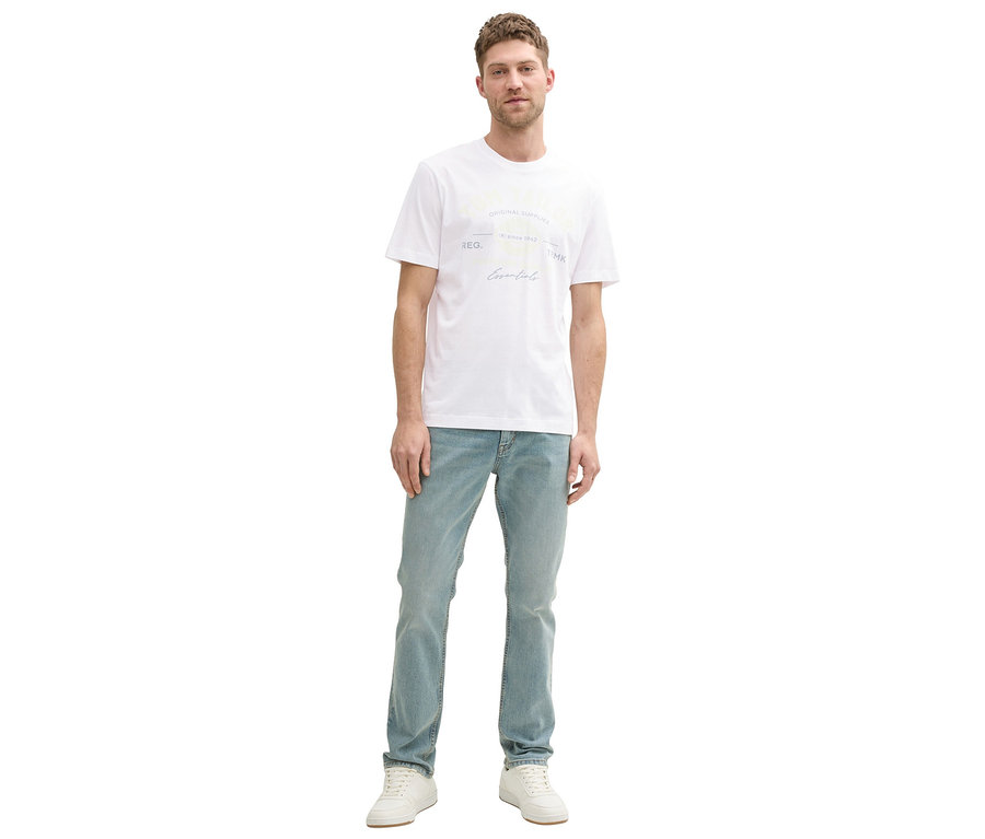 Mężczyzna stoi w jeansach i białej koszulce z logo Tom Tailor.