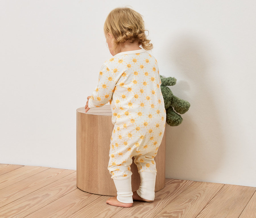 Małe dziecko w żółto-białym kombinezonie w słoneczka stoi obok okrągłego drewnianego stołka, obok znajduje się zielona pluszowa zabawka.