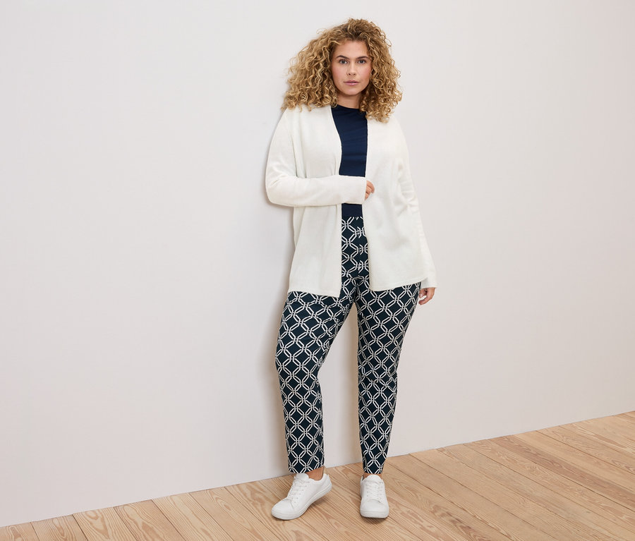 Kobieta o kręconych włosach w białym kardiganie, granatowej bluzce i wzorzystych spodniach na tle białej ściany.