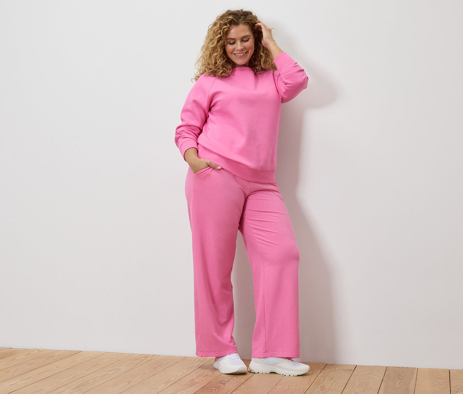 Kobieta z kręconymi włosami w różowym dresie i białych tenisówkach, trzymająca rękę na głowie.