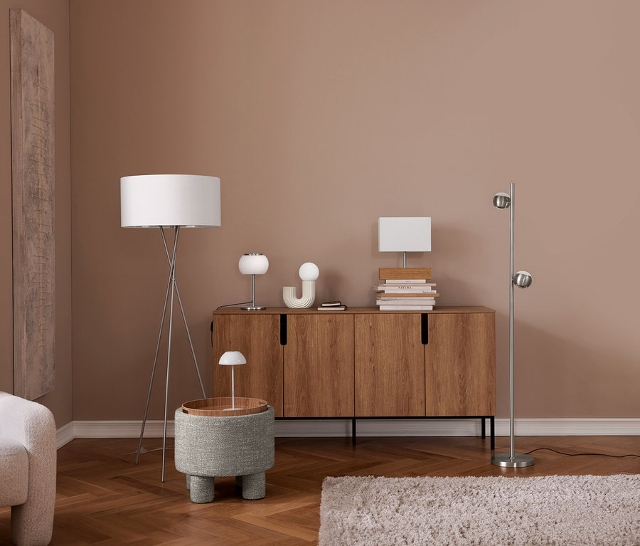 Nowoczesny salon z jasnobrązowymi ścianami i drewnianą podłogą. Po lewej kremowy fotel. Lampa podłogowa na trójnogu stoi obok szarego pufa z małą lampką. Drewniana komoda mieści dwie lampy stołowe, przedmiot dekoracyjny i książki. Po prawej srebrna lampa podłogowa z dwoma kulistymi światłami, na podłodze jasny dywan.
