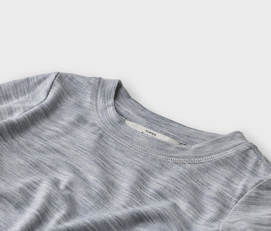 Zbliżenie szarego melanżowego T-shirtu z okrągłym dekoltem, widocznymi białymi szwami wokół kołnierza i białą metką z napisem „TCHIBO”.