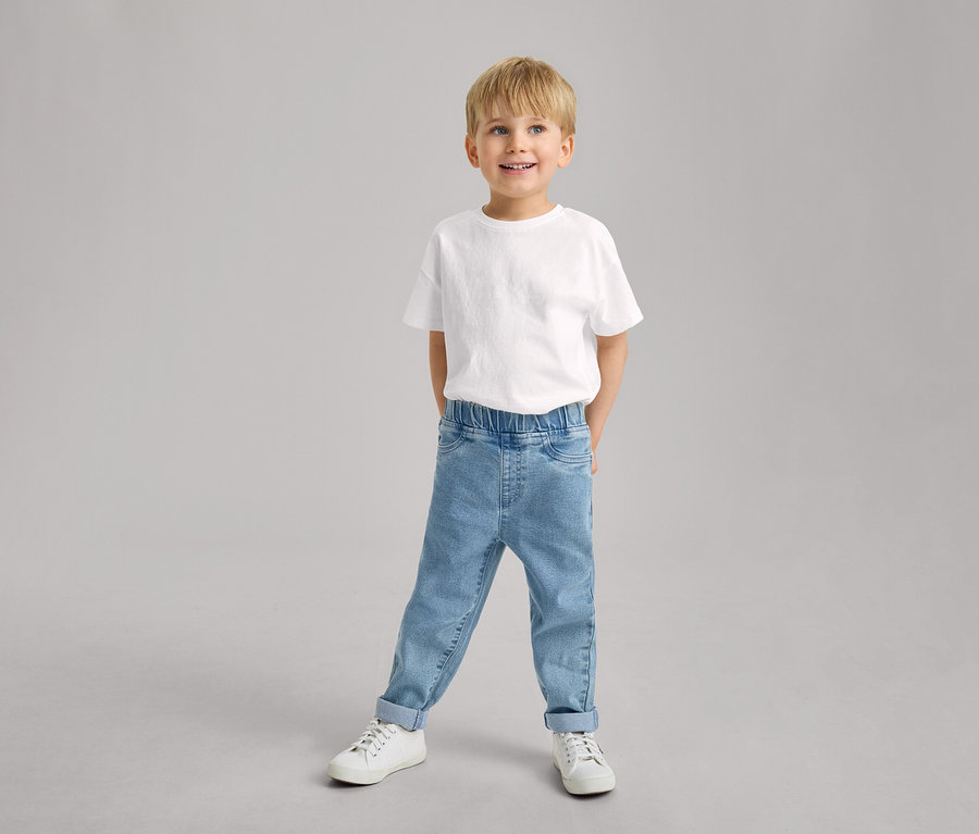 Uśmiechnięty mały chłopiec stoi w białym T-shircie, jasnoniebieskich spodniach na gumce – fason „Luca” z podwiniętymi nogawkami i białych tenisówkach.