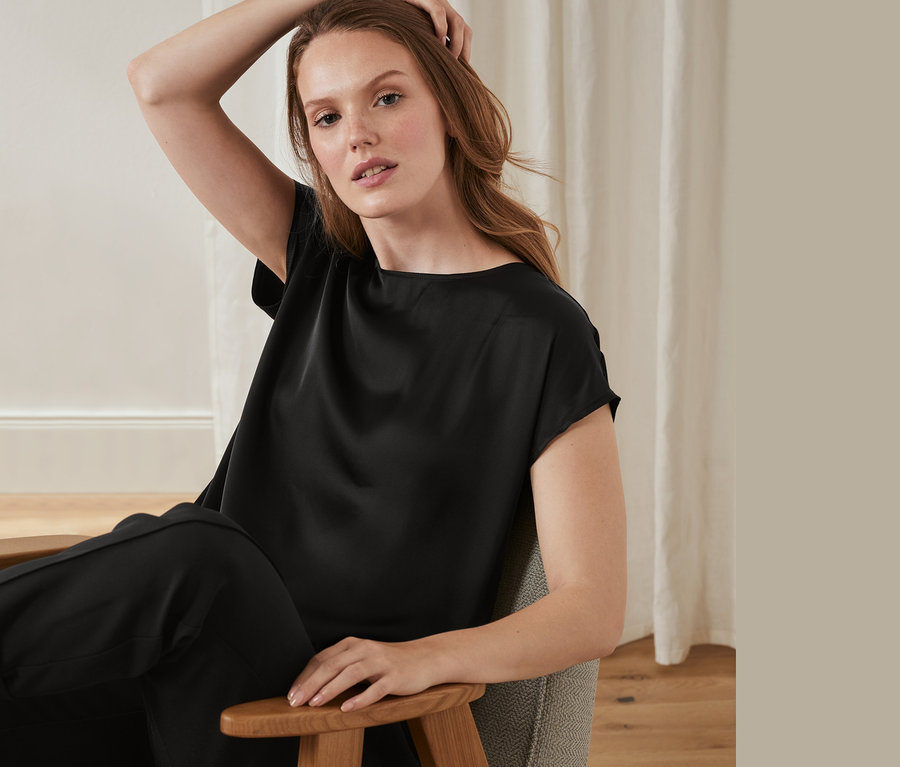 Kobieta siedzi w fotelu, ubrana w bluzkę z satynowym przodem.