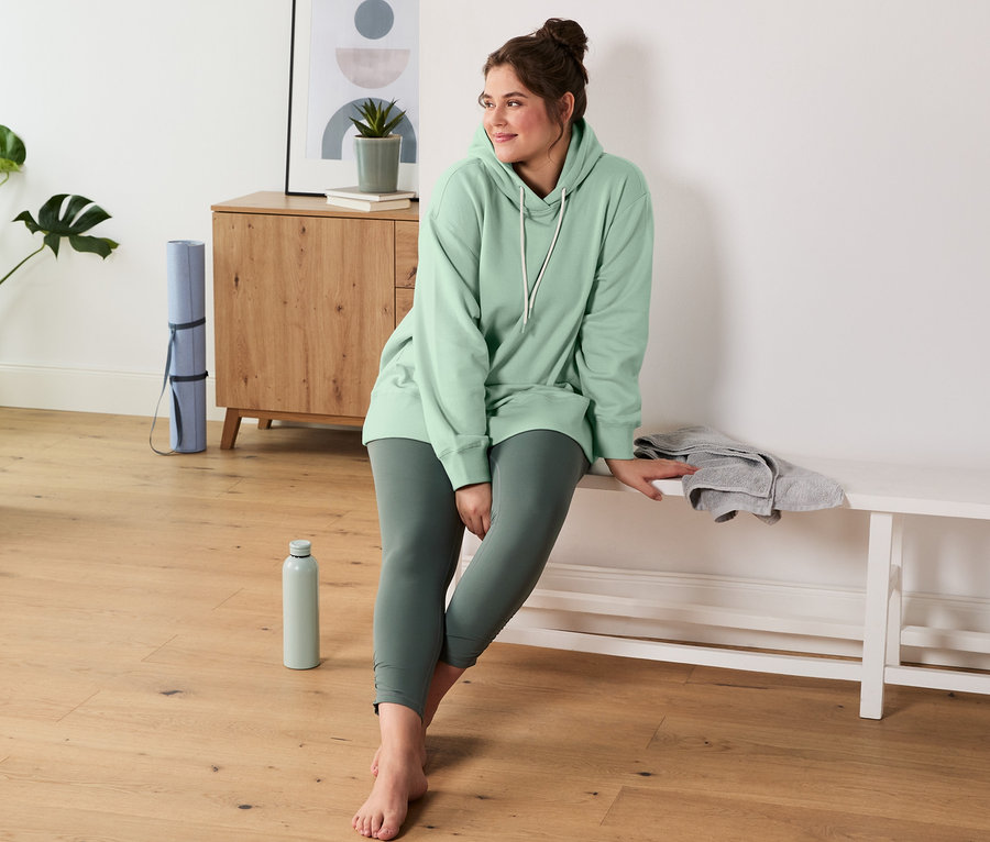 Kobieta siedzi na ławce, ubrana w zieloną bluzę dresową z kapturem i legginsy sportowe modelujące sylwetkę.