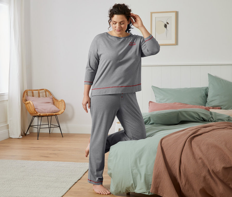 Kobieta w szarej piżamie w delikatne paski stoi przy łóżku.