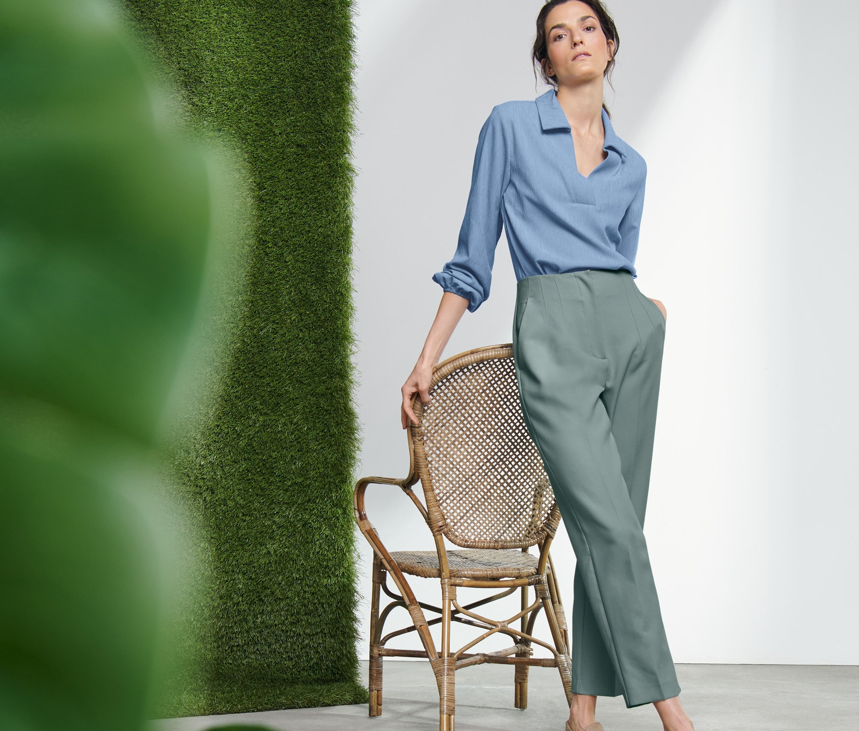 Kobieta pozuje obok wiklinowego krzesła, ubrana w spodnie z tkaniny.
