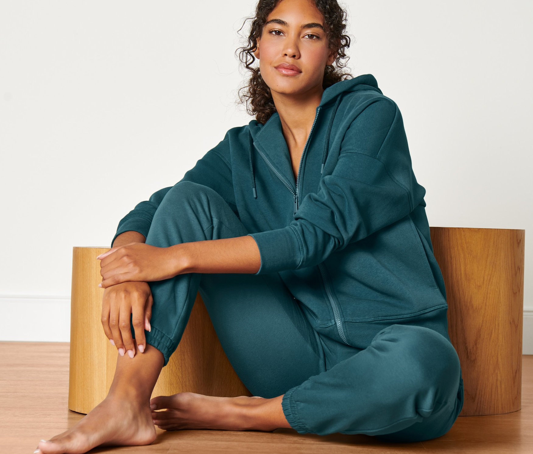Kobieta siedzi na drewnianym stołku, ubrana w bluzę dresową w kolorze ciemnej zieleni szmaragdowej.