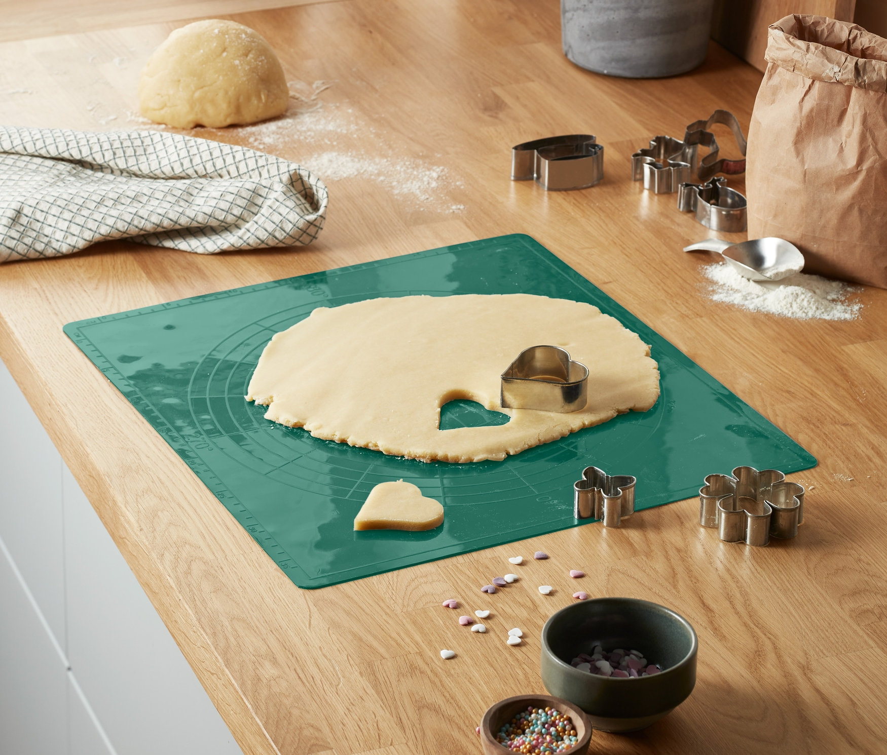 Na blacie kuchennym leży zielona mata do pieczenia z rozwałkowanym ciastem i foremkami do ciastek.