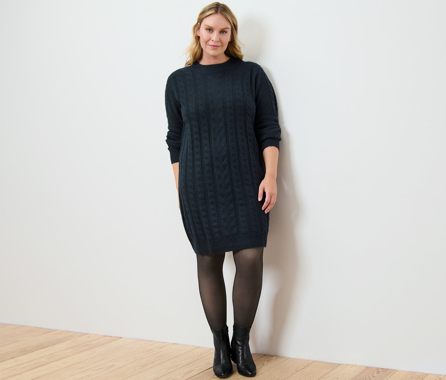 Kobieta pozuje w sukience z dzianiny z warkoczowym wzorem i cienkich rajstopach 50 DEN z efektem przezroczystości.