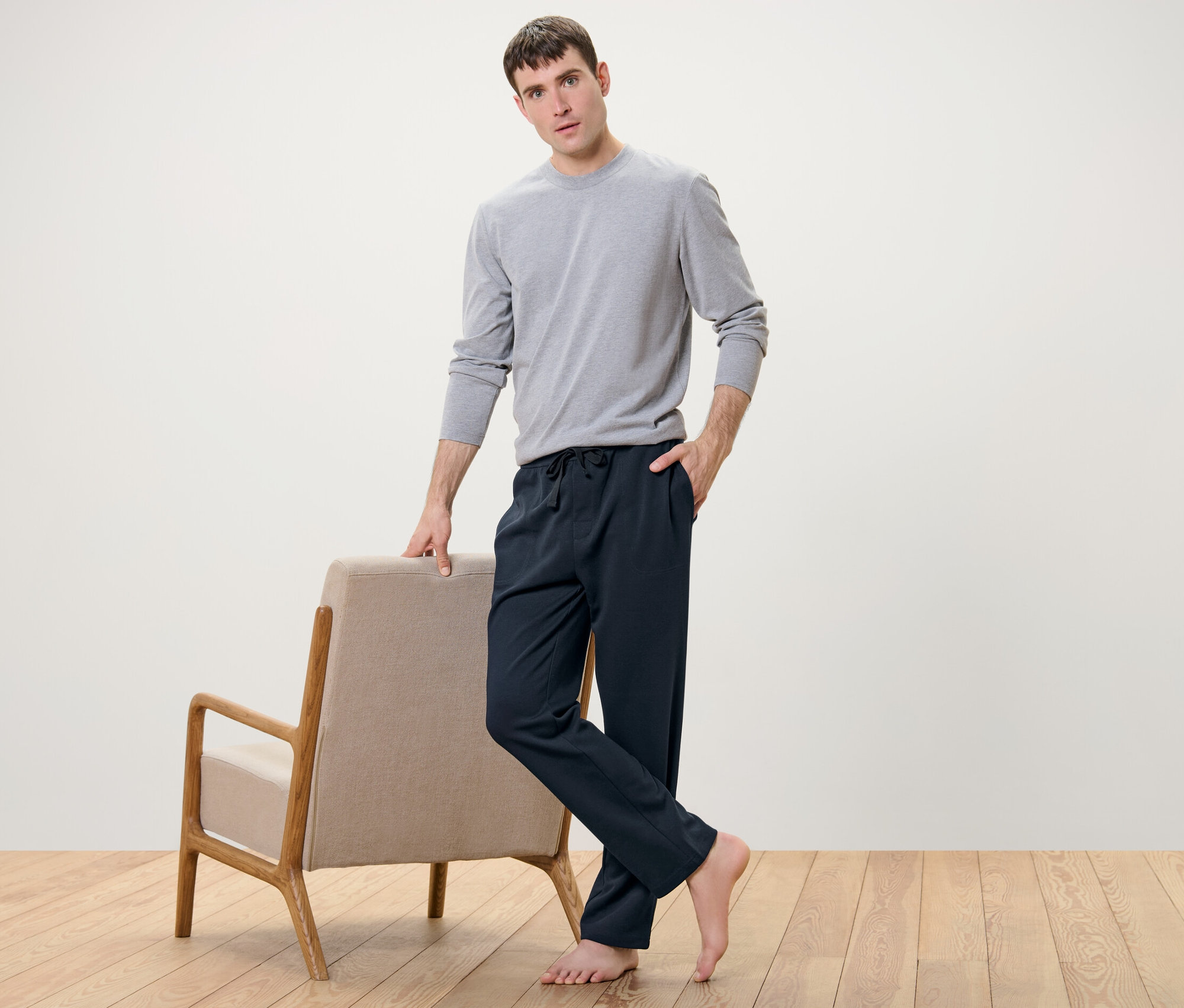 Mężczyzna pozuje obok fotela, ubrany w spodnie rekreacyjne i szarą koszulę.