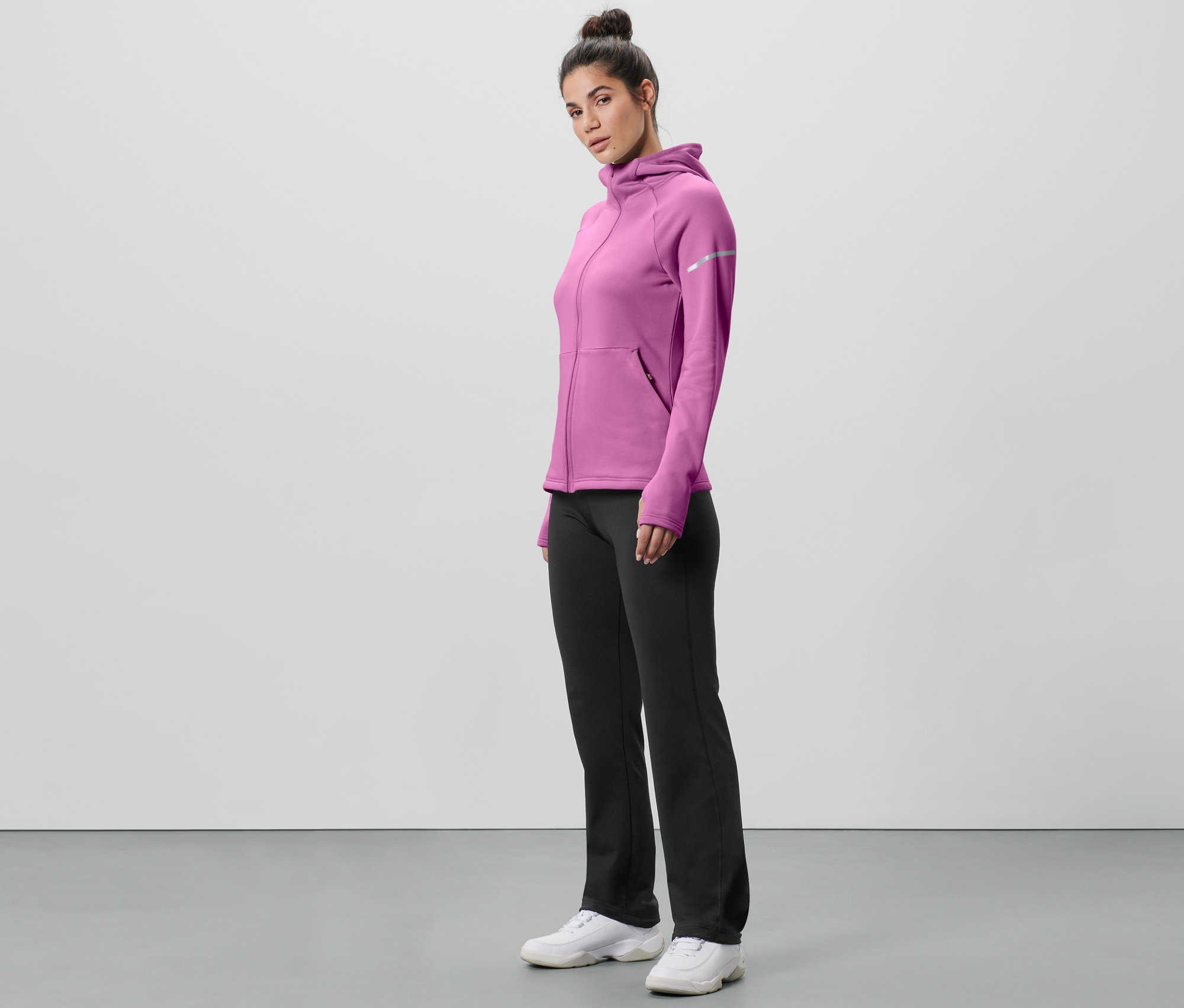 Kobieta stoi bokiem w różowej bluzie z kapturem i czarnych spodniach termoaktywnych.