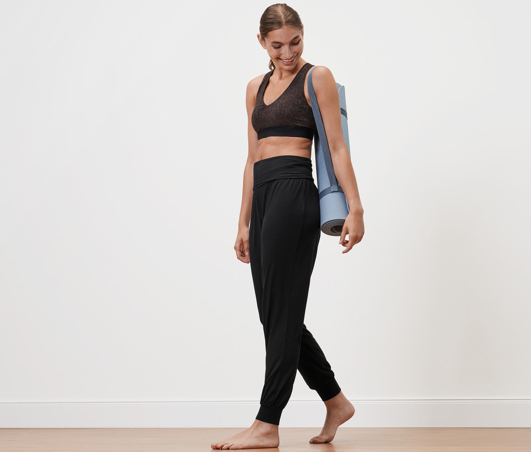 Kobieta idzie boso z matą do jogi na ramieniu, ubrana w czarne spodnie do jogi.