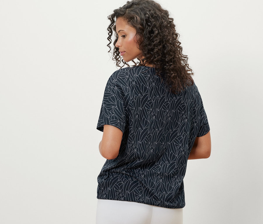 Widok z tyłu kobiety z kręconymi włosami, ubranej w granatową bluzkę z obniżoną linią ramion i białe spodnie.