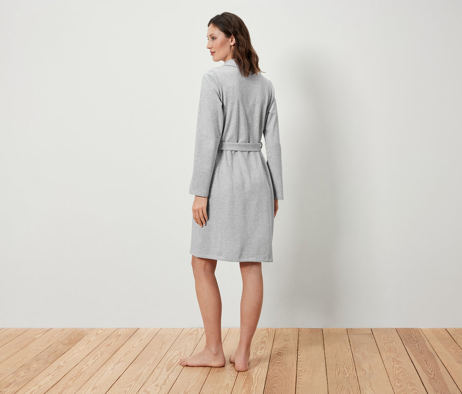 Kobieta stoi tyłem, ubrana w szary szlafrok.